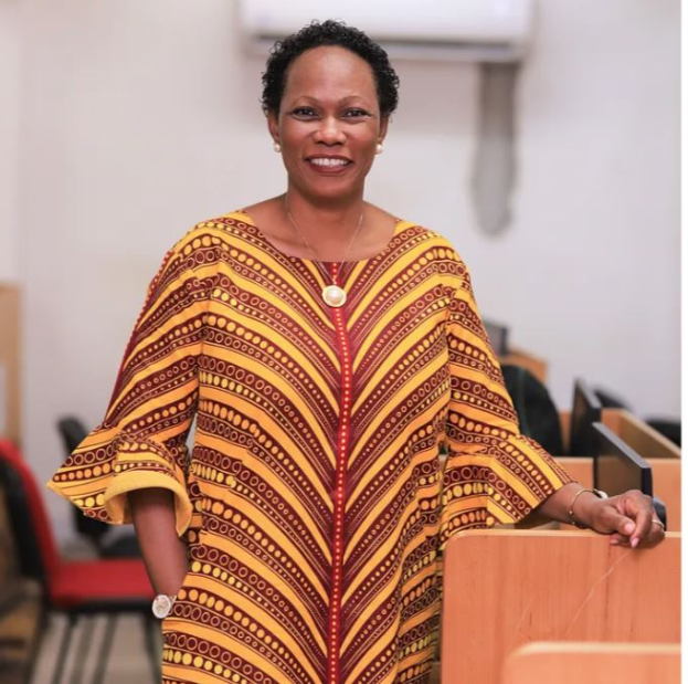 Eine Frau in einem gelben und roten Kleid steht an einem Pult mit einem Mikrofon, spricht in einer öffentlichen Versammlung mit Stühlen und einer Klimaanlage im Hintergrund.