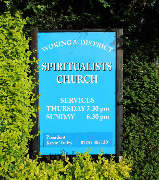 Ein Holzbrett mit der Aufschrift "Woking & District Spiritualists Church" vor Pflanzen, Bäumen und einem Gebäude im Hintergrund.