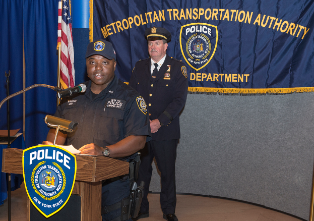 Zwei Polizeibeamte stehen vor einem Rednerpult, einer Tafel mit Symbolen und Text, einer Fahne, einem Stand und einem Tisch, wobei einer der Beamten Papiere hält und in ein Mikrofon spricht.
