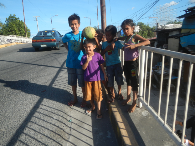Eine Gruppe von Kindern steht an der Straße, einer hält einen Ball in der Hand, rechts ist ein Geländer zu sehen, im Hintergrund Fahrzeuge, Strommasten, Laternen, Bäume und ein bewölkter Himmel.