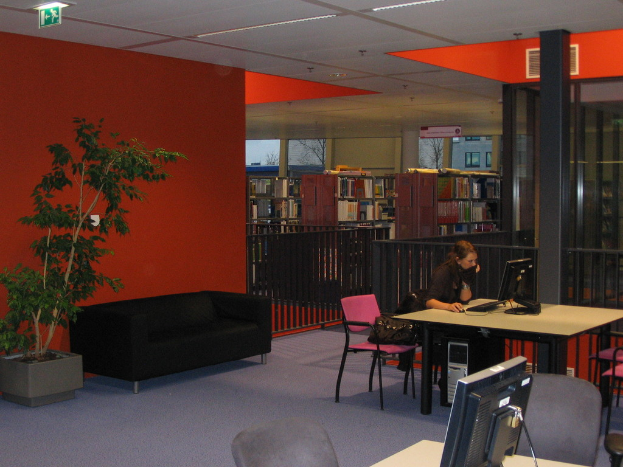 Eine Frau in einem schwarzen T-Shirt bedient ein Computersystem auf einem Tisch, mit einer schwarzen Couch dahinter, einer roten Wand mit einer Pflanze und Bücherregalen im Hintergrund, alles innerhalb eines Raums mit einer weißen Wand, Fenstern und einer sichtbaren Decke.