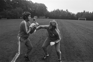 Zwei Männer in Schwarz-Weiß-Uniformen sind in eine körperliche Auseinandersetzung auf einem Fußballfeld verwickelt, mit Bäumen und einer Hütte im Hintergrund.