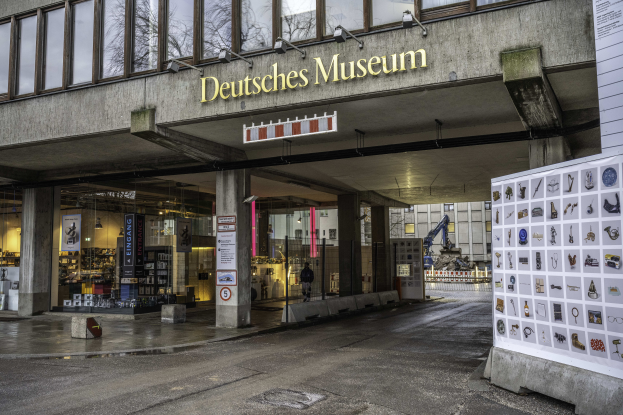 Das Deutsche Museum in Berlin, Deutschland, ein großes Gebäude mit Glasfenstern, Säulen und einer Namensschild, mit einer Tafel mit Bildern und Text auf der rechten Seite und anderen Gebäuden, Bäumen und dem Himmel im Hintergrund.