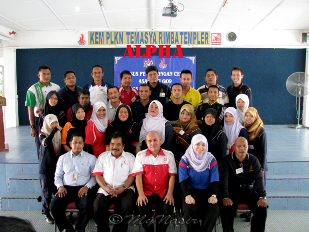 Eine Menschenmenge, die vor einer Bühne strahlt und posiert, mit einer Wandtafel dahinter, einem Tischventilator rechts, einem Podium links und einem Projektor oben im Bild.