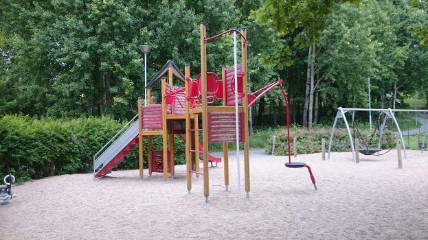 Ein Spielplatz mit einer roten und gelben Spielstruktur in der Mitte, umgeben von Sand, Schaukeln, einer Rutsche, einem Lichtmast, Pflanzen und Bäumen unter einem klaren blauen Himmel.