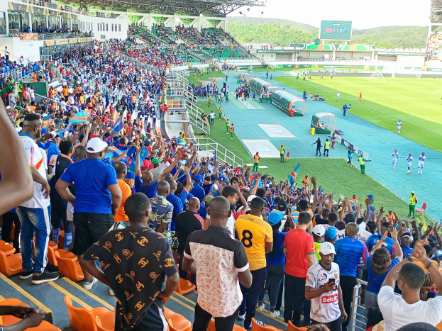 Eine große Menschenmenge versammelt sich vor einem eingezäunten Fußballfeld mit Hügeln und einem klaren blauen Himmel und erwartet den Beginn eines Spiels während der African Cup of Nations 2019.