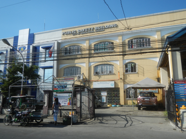 Ein Gebäude mit einem Schild "Feuerwehrhaus" umgeben von Bäumen, Straßenlaternen, Straßenlaternen, Fahrzeugen, Fußgängern, Gittern, Namensschildern, Strommasten und Kabel unter einem klaren blauen Himmel.