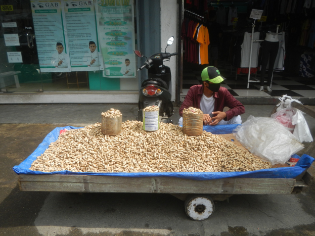 Ein Mann in einer Mütze sitzt auf einem Erdnusswagen mit einem Roller in der Nähe, während Kleidung im Hintergrund hängt und ein Geschäft mit Plakaten zu sehen ist.