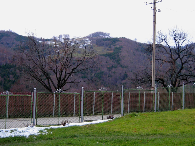Eine grüne Landschaft mit einem Zaun, einem Pfahl und Bäumen, mit ein paar Häusern im Hintergrund auf einem Hügel.