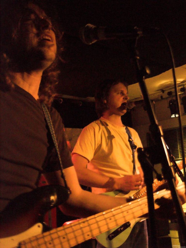 Eine Person in einem wei├čen Hemd singt in ein Mikrofon und spielt eine grüne Gitarre, steht neben einer anderen Person in einem schwarzen Hemd und Brille, die ebenfalls singt und eine schwarze Gitarre spielt, mit Scheinwerfern und Musikinstrumenten im Hintergrund.