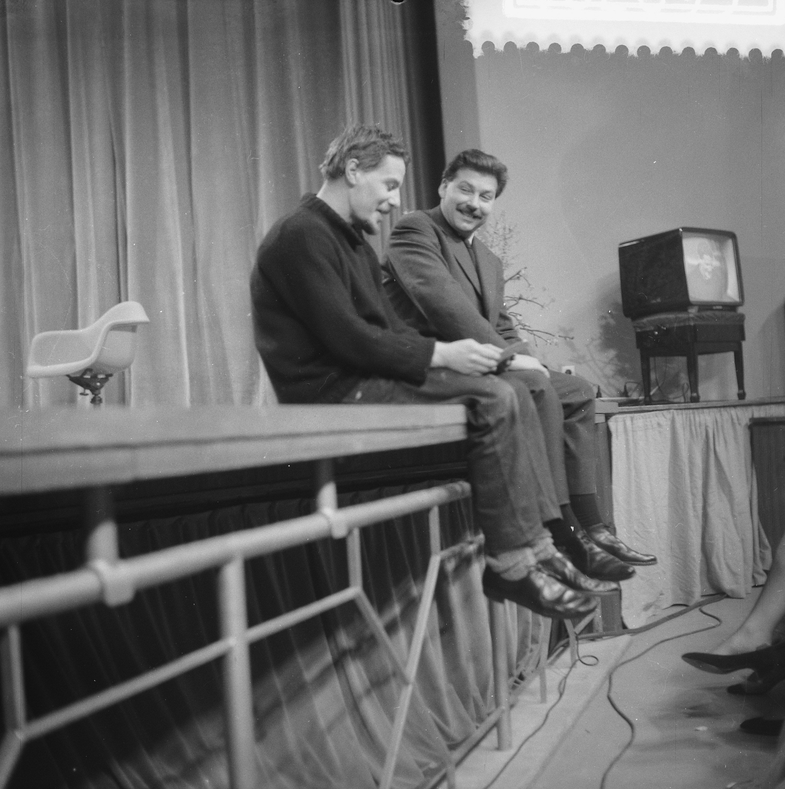 Zwei Männer sitzen auf einer Bühne, sie lächeln, rechts eine Person auf dem Boden, links ein leerer Stuhl und Vorhänge und eine Wand im Hintergrund.