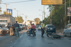 Eine Gruppe von Menschen um ein verunglücktes Motorrad auf der Stra√enseite mit mehreren Fahrzeugen, darunter ein LKW, und einem Hintergrund aus Bäumen, Masten, Lichtern und Schildern unter dem Himmel.