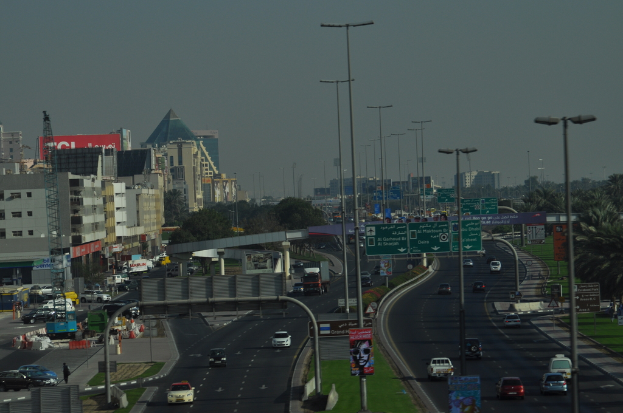 Eine Luftaufnahme einer Stadt mit Gebäuden, Türmen, Fahrzeugen, Werbetafeln, Schildern, Straßen, Bäumen, Straßenlaternen, Menschen, Absperrgittern und Informationsschildern unter einem klaren Himmel.