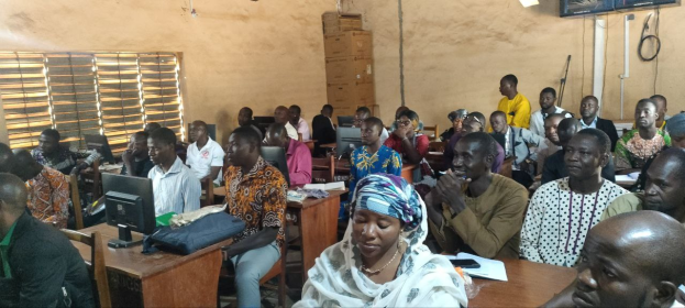 Eine Gruppe von Menschen sitzt an Tischen in einem Klassenzimmer mit Monitoren, Taschen, Büchern und Papieren und nimmt an einem Techniktraining teil; ein Fenster links, Kartons und ein Fernseher an der Wand im Hintergrund.