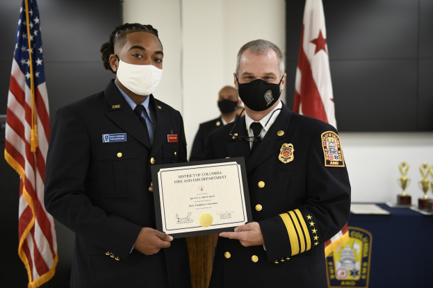 Zwei uniformierte Personen, der District of Columbia Fire Chief links und der Assistant Fire Chief rechts, stehen nebeneinander und tragen Masken sowie ein Zertifikat, mit einem Awards-Tisch und Fahnen im Hintergrund.