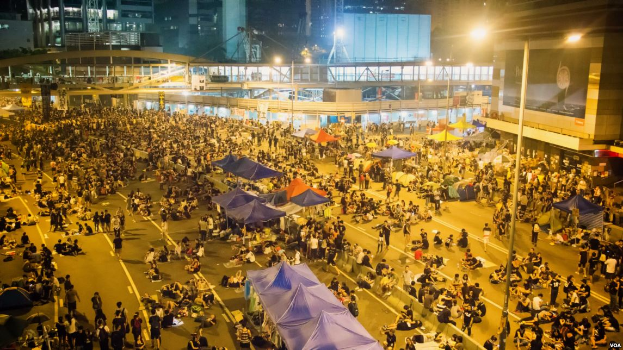 Eine große Menschenmenge steht nachts auf einer Straße, beleuchtet von Straßenlaternen, umgeben von gläsernen Gebäuden, mit Zelten, Pfählen und einer Brücke mit Geländern im Hintergrund, wahrscheinlich protestierend in Hong Kong gegen das Auslieferungsgesetz.