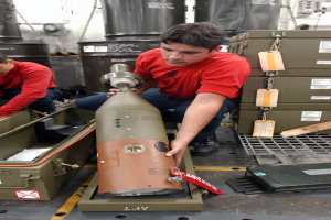 Zwei Männer in roten Hemden arbeiten an einem großen Metallfragmentationssprengkörper, mit Kisten, Ketten und anderen Gegenständen im Hintergrund.