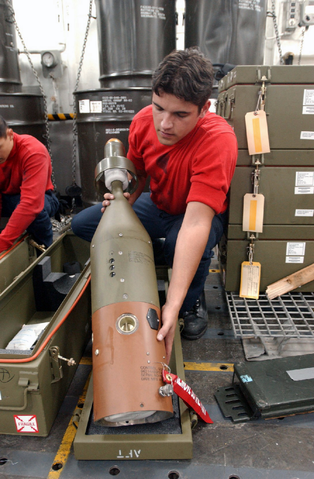 Zwei Männer in roten Hemden arbeiten an einem großen Metallfragmentationssprengkörper, mit Kisten, Ketten und anderen Gegenständen im Hintergrund.