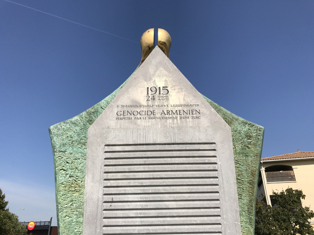 Ein hoher Denkmal mit einer Uhr darauf steht in einem Park, umgeben von Bäumen und Gebäuden unter einem klaren Himmel, mit eingravierten Texten, die den 24. Jahrestag des Völkermords in Armenien gedenken.