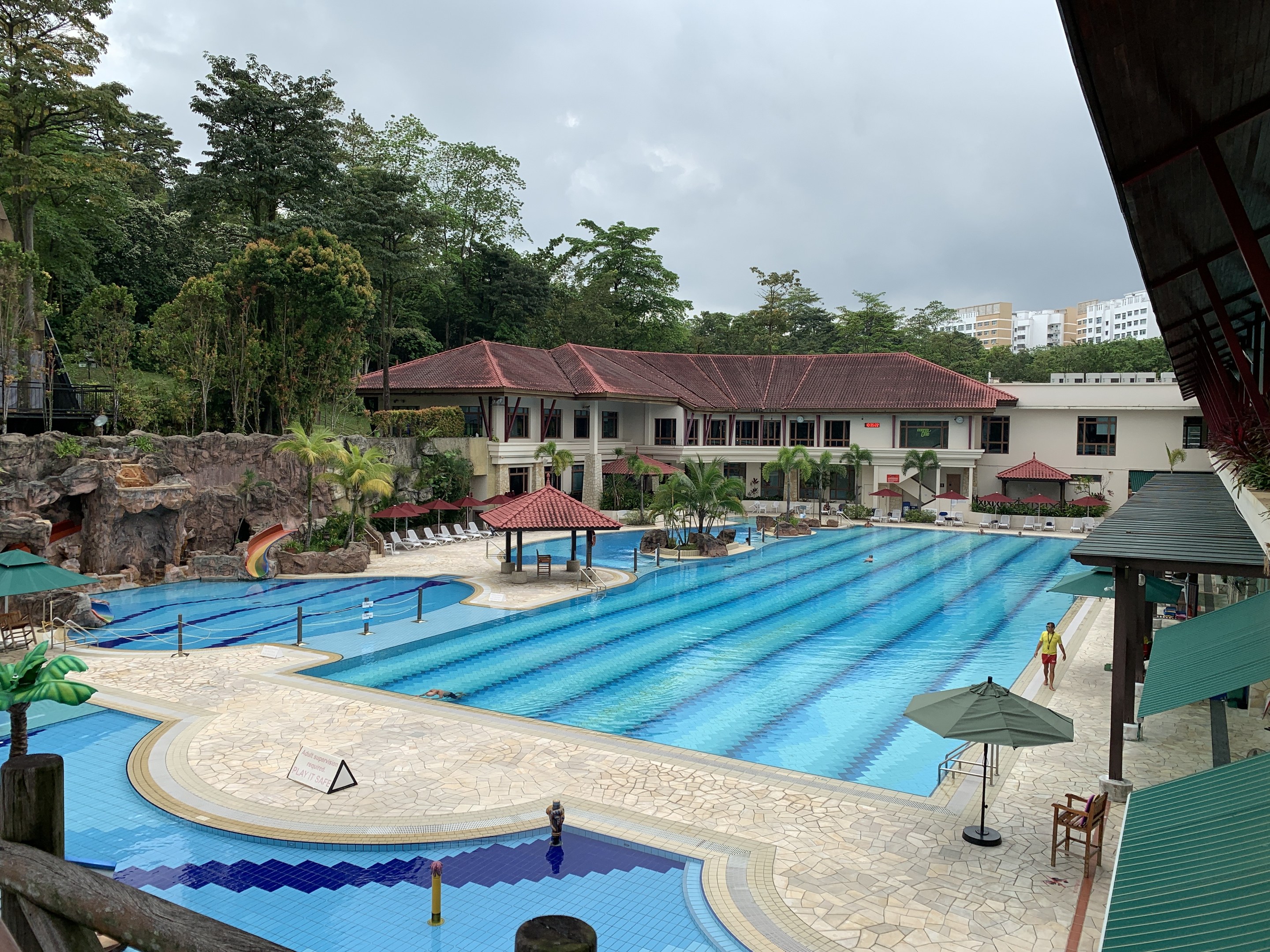 Großes Schwimmbad im Ritz-Carlton, Kota Kinabalu, mit Menschen, Sonnenschirmen, Liegestühlen, Tischen, Grünpflanzen, Felsen, Gebäuden und einem Schuppen auf der rechten Seite sowie einem sichtbaren Himmel im Hintergrund.