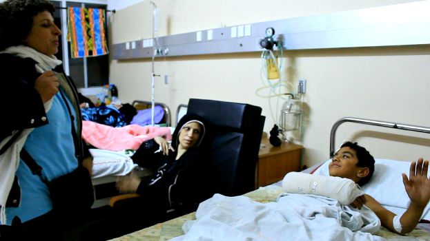 Einblicke in ein Krankenhaus mit Betten und Patienten, Kochsalzflaschen auf Ständern zwischen den Betten und einer Frau mit einer Tasche in der Nähe eines Patienten.