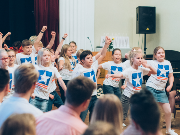 Eine Gruppe von Kindern in weißen Hemden und blauen Shorts, die vor einem sitzenden Publikum tanzen, mit einer Vorhangwand, Lautsprecherstativ und einem Tisch mit Mikrofon im Hintergrund.