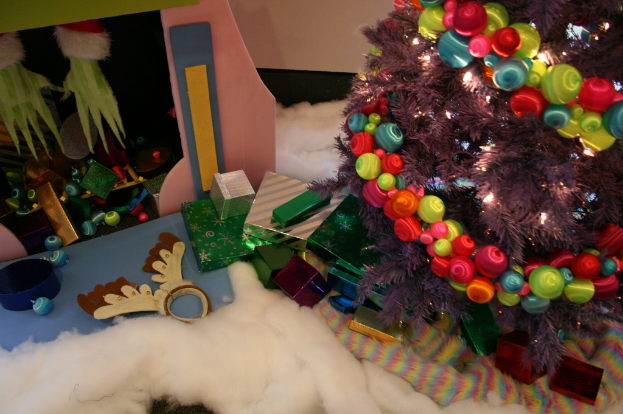 Drinnen steht ein geschm├╝ckter Weihnachtsbaum mit Lichterkette und Schmuck rechts, Geschenkboxen und Deko in der Mitte, eine H├╝tte mit Deko oben links und Baumwolle und Wolle unten.