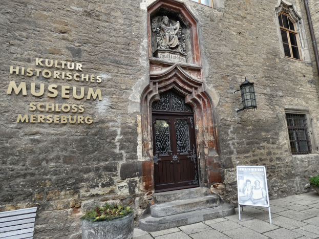 Das Kulturhistorische Museum in Schloss Merseburg, Deutschland, ein Gebäude mit Fenstern, einer Tür, einem Schild, einer Bank, Topfpflanzen und einer Pfeife unter einem bewölkten Himmel.