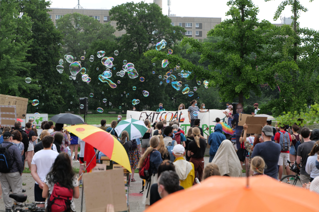 Eine Gruppe von Menschen, die vor einer Menge mit Schirmen, Taschen und Schildern mit Text steht, mit Bäumen, Gebäuden, einem Turm und einem klaren blauen Himmel im Hintergrund, einige Menge-Mitglieder Seifenblasen pusten.