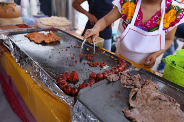 Eine Frau in einer Schürze grillt auf einem Markt, mit einem Tisch voller Lebensmittel, einem Eimer und anderen Gegenständen in der Nähe und einigen Menschen im Hintergrund.