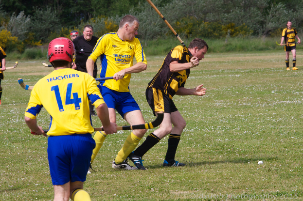 Menschen spielen Hockey auf einer grünen Wiese mit anderen und Grünflächen im Hintergrund.