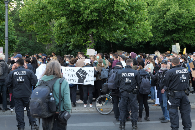 Eine große Gruppe von Menschen nimmt an einer Black Lives Matter Demonstration in Berlin teil, einige halten Schilder, andere tragen Mützen und Taschen, mit einem Fahrrad im Vordergrund und Bäumen und einem Pfahl im Hintergrund.