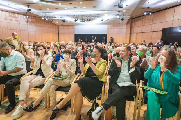 Eine Gruppe von Menschen in Stühlen klatschend, einige tragen Masken, mit Taschen auf dem Boden, vor einer Menge bei einer Coronavirus-Konferenz mit einem Bildschirm und Deckenleuchten im Hintergrund.