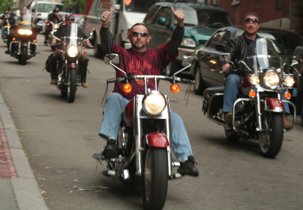 Eine Gruppe von Menschen, die auf Fahrrädern auf einer Straße fahren, wobei eine Person in einem roten Hemd ohne Hände auf dem Lenker und eine andere Person in einer schwarzen Jacke lacht. Im Hintergrund sind zwei Autos und ein Lieferwagen zu sehen.