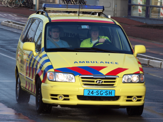 Ein gelber Krankenwagen mit zwei sichtbaren Insassen fährt an einem Gebäude mit Glasfenstern vorbei, während Fahrräder auf dem benachbarten Gehweg abgestellt sind.