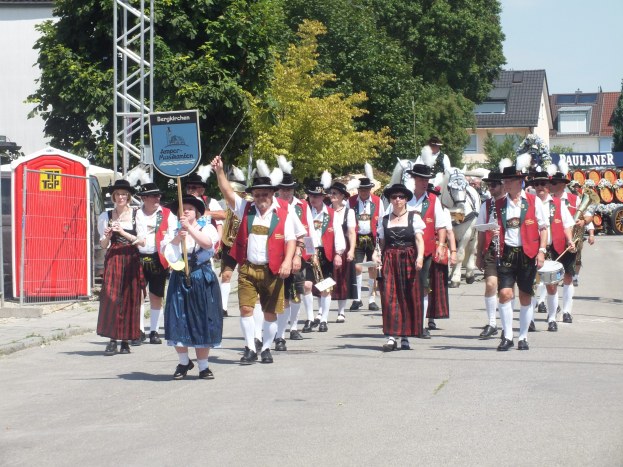 Eine Gruppe von Menschen in traditioneller bayrischer Tracht, die eine Straße entlanggehen, einige halten Musikinstrumente und tragen Hüte, mit Bäumen, Gebäuden und einem klaren blauen Himmel im Hintergrund; ein rotes Objekt und eine Tafel mit Text sind links sichtbar.