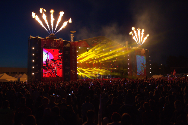 Eine große Menschenmenge auf einem Musikfestival mit beleuchteter Bühne, Performern, Bannern, Zelten links und Feuerwerk am dunklen Himmel.