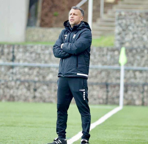 Ein Füßballtrainer, der auf einem Feld steht und die Ärmel überkreuzt, mit einer schwarzen Jacke und Hose, vor einem Zaun, einer Treppe und einer Wand.