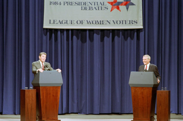Zwei Männer in Blazern und Krawatten stehen an Podien mit Mikrofonen und Gläsern vor einem blauen Vorhang und einem Banner mit Text und Sternen, diskutieren die Präsidentschaftswahldebatten von 1984.