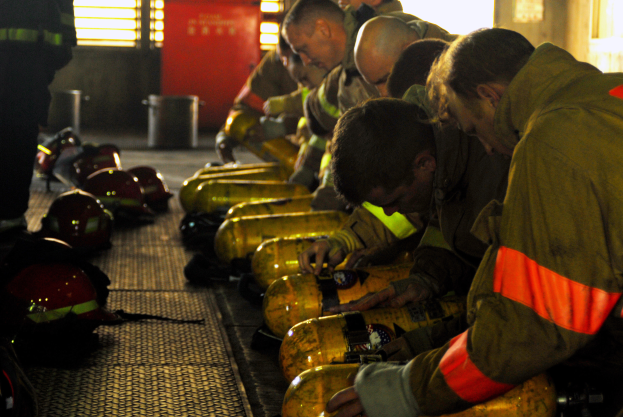 Eine Gruppe von Feuerwehrleuten arbeitet an einem Feuerwehrauto umgeben von verstreuten Helmen und Zylindern, mit einer Wand aus Fenstern und einer Tür im Hintergrund.