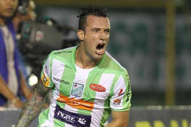 Ein Fussballspieler in grünem und weißem Trikot mit Tattoos auf dem Arm feiert nach einem Tor, mit unscharfen Figuren und einer Tafel im Hintergrund.