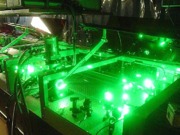 Eine Lasermaschine auf einem Tisch, die ein grünes Licht ausstrahlt, mit sichtbaren Komponenten und verbundenen Kabeln.