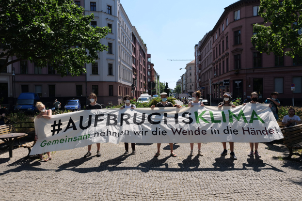 Eine Gruppe maskierter Personen hält ein Transparent mit der Aufschrift 'Aufbruchsklima' und protestiert gegen den Klimawandel in Berlin, Deutschland, mit Gebäuden, Bänken, Tischen, Pflanzen, Bäumen, Fahrzeugen, Laternenmasten und Schildern im Hintergrund.