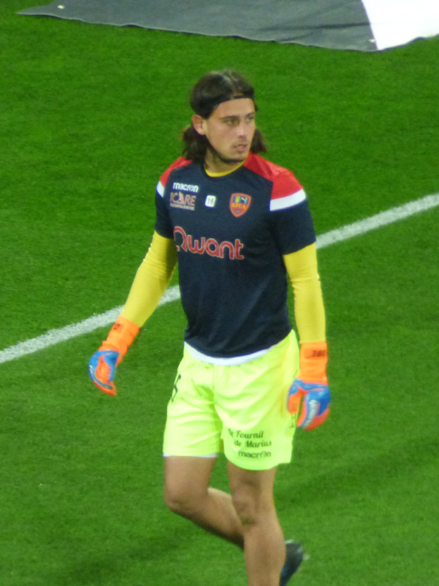 Ein Mann in einem blauen Hemd und gelben Shorts läuft auf einem Fußballfeld mit ausgestreckten Armen und entschlossener Miene, mit einem Tuch oben auf dem Bild.