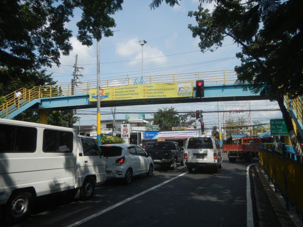 Eine belebte Straße mit Fahrzeugen, eine Brücke mit Geländern und Treppen, Laternen, Verkehrsampeln, Informationsschilder, Bäume, Gebäude und ein bewölkter Himmel.