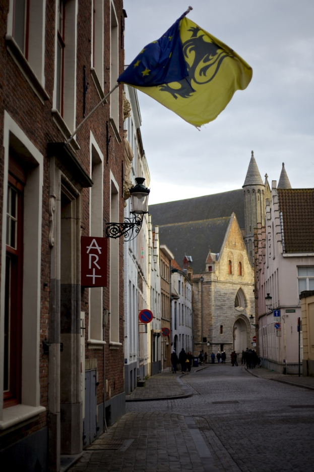 Eine Stadtansicht mit Gebäuden, Menschen auf der Straße, Pfählen, Schildern und einer Flagge an einem der Gebäude, mit dem Himmel oben und der Straße unten.