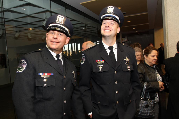 Zwei grinsende Beamte im Vordergrund, mit Menschen in der Mitte, Glasfenstern links, Lampen und Decke oben und Pfeilern und mehr Menschen rechts.