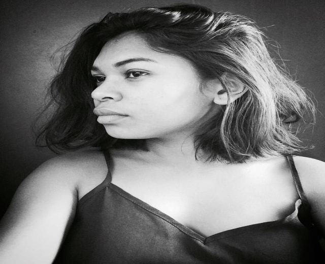 Eine selbstbewusste Frau in einem schwarzen Kleid posiert in einem Schwarz-Weiß-Foto vor einer Wand mit einem glatten Bob und natürlichem Make-up.