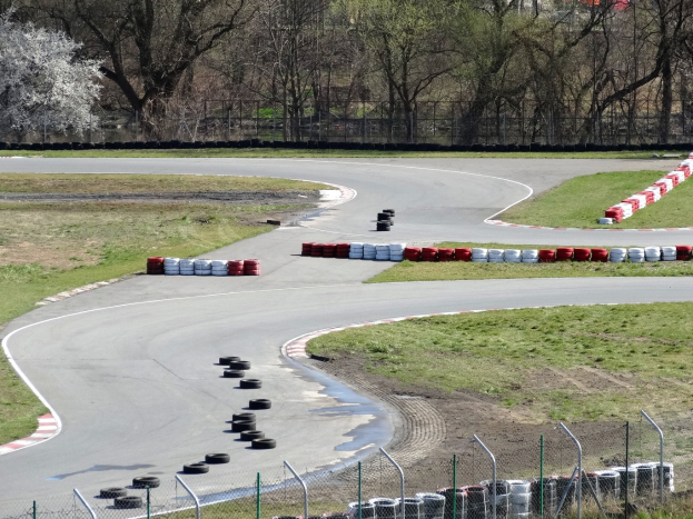 Rennstrecke mit zahlreichen Reifen, die an ihren Rändern angeordnet sind und von Gras und Maschendrahtzaun umgeben ist, mit Bäumen im Hintergrund.