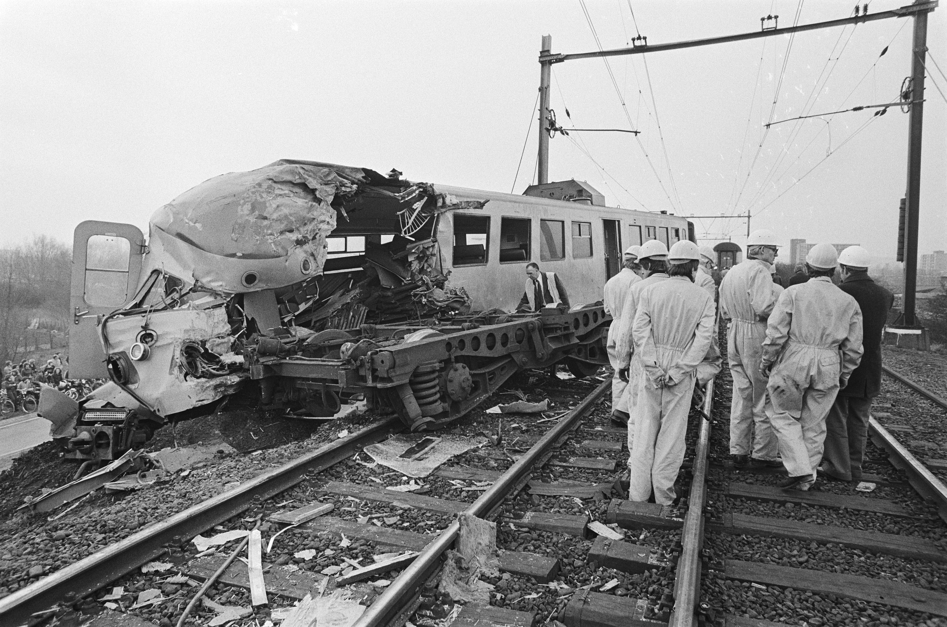 Ein Schwarz-Weiß-Bild einer Zugkollision auf den Schienen mit Menschen in Helmen in der Nähe, umgeben von Strommasten, Bäumen, Gebäuden und einem klaren Himmel.